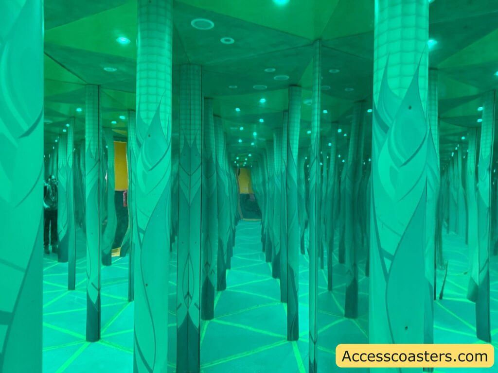 Mirror maze with green lighting. The image shows numerous vertical columns with leaf-like patterns, reflected infinitely by mirrors on the walls and possibly on the floor, creating a confusing and disorienting visual effect. The ceiling has small round lights, and the floor has yellow lines forming geometric shapes.