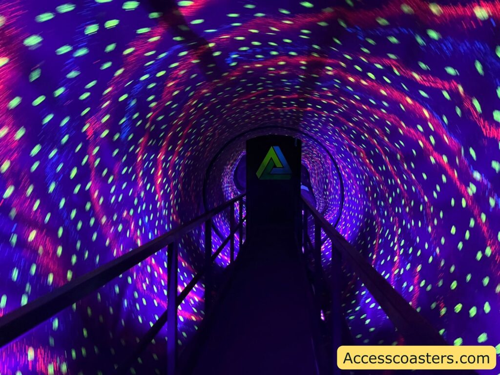 Tunnel illusion attraction with a walkway, surrounded by swirling, colorful neon lights in purple, blue, green, and pink. The lights create a spinning vortex effect on the tunnel walls, giving a sense of motion and disorientation. At the end of the walkway, there is a door with a green triangular logo.