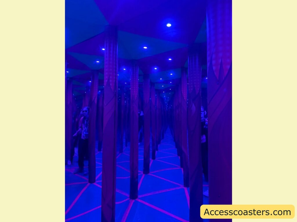 Mirror maze with blue and purple lighting. The image shows a series of tall, vertical columns and mirrors creating a repeating, endless effect. The floor has intersecting red lines, and the ceiling is illuminated with small blue lights. A person is partially visible on the left, reflected multiple times in the mirrors.
