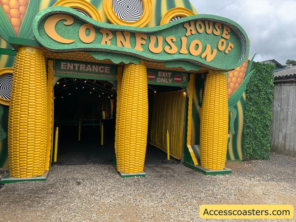 Amusement attraction entrance themed like corn, with large yellow corn cobs framing two doorways. The sign above reads "House of Cornfusion" in playful, wavy letters. The left doorway is labeled "Entrance" and the right "Exit Only." The building is decorated with green and yellow corn imagery,