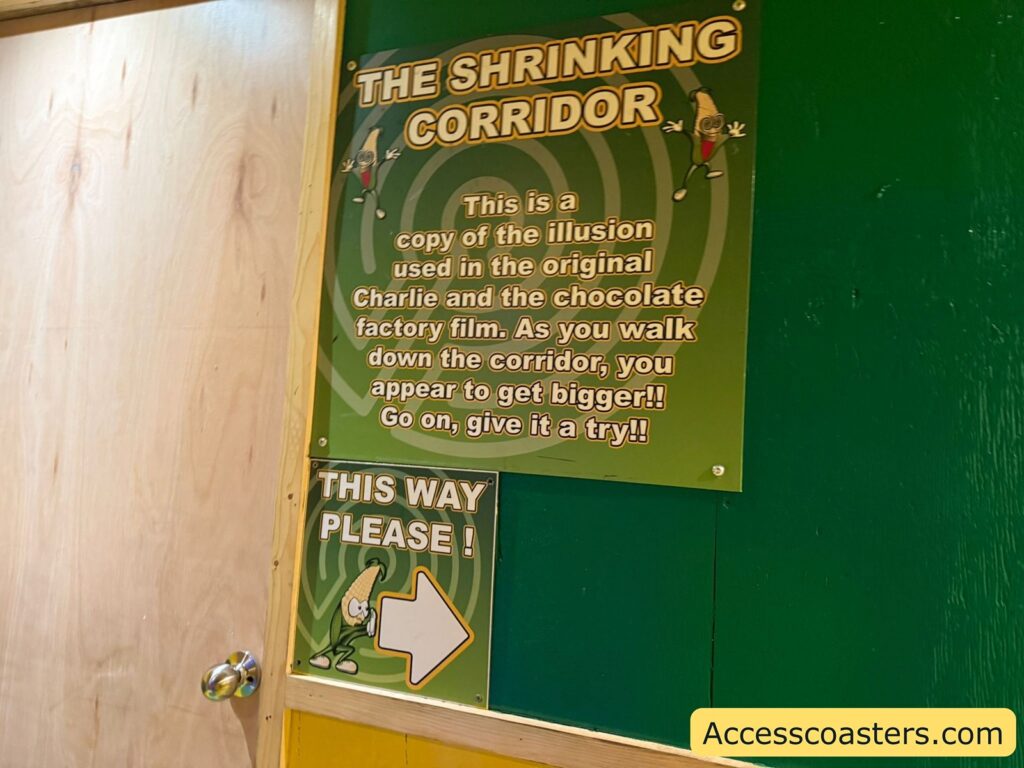 Signage on a green and yellow wall. The largest sign reads: "THE SHRINKING CORRIDOR. This is a copy of the illusion used in the original Charlie and the Chocolate Factory film. As you walk down the corridor, you appear to get bigger!! Go on, give it a try!!" The sign has a green spiral background and cartoon corn characters on either side.

Below it, another sign says: "THIS WAY PLEASE!" with a large white arrow pointing right and another cartoon corn character.