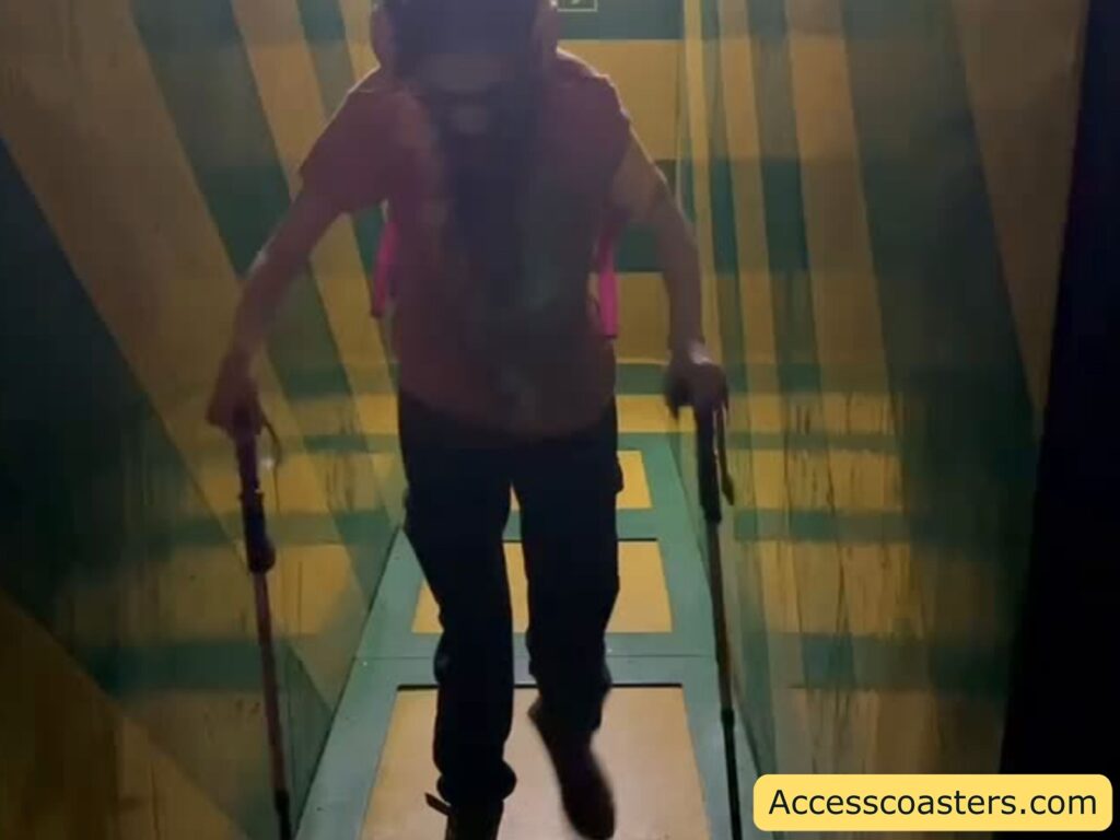 A person using two walking sticks is moving through a narrow, slanted corridor with bold yellow and green stripes on the walls and floor. The lighting is dim, casting shadows. 