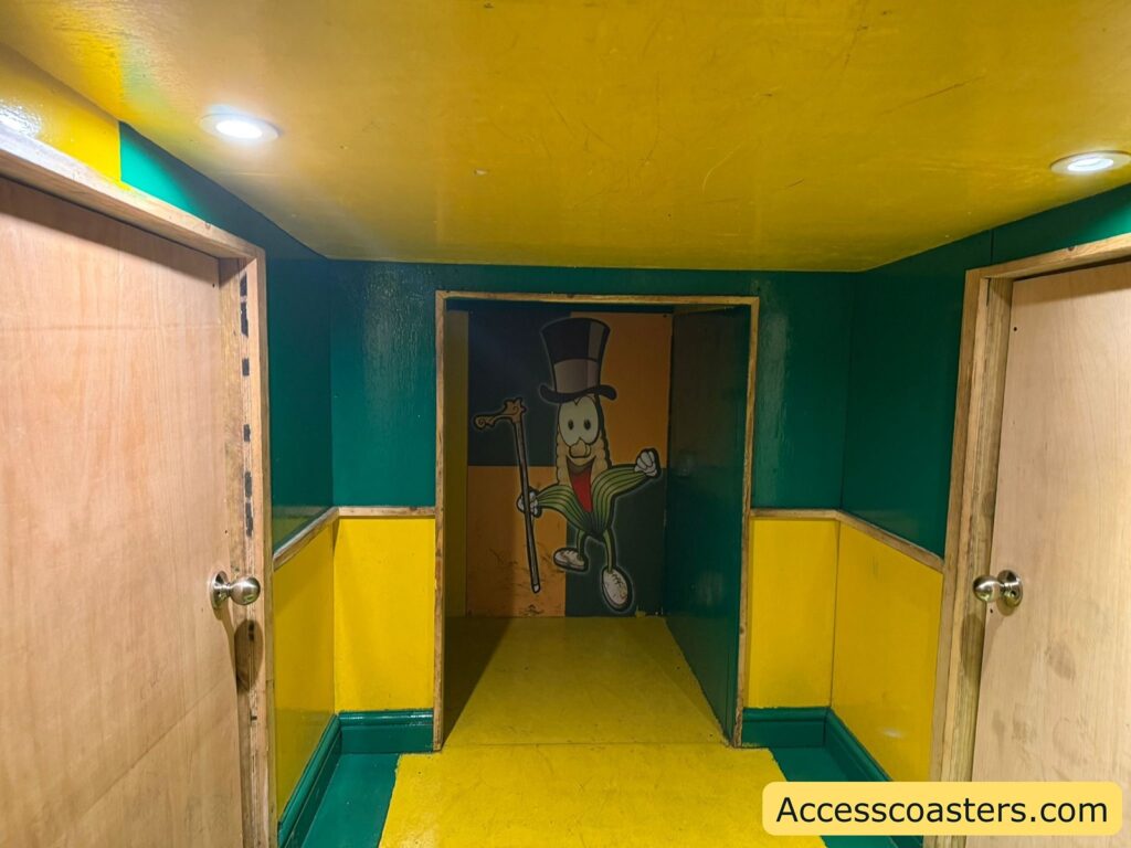 A brightly colored hallway with yellow and green walls and ceiling, illuminated by recessed lights. There are two wooden doors on either side with silver knobs. At the end of the hallway, there is a cartoon mural of a smiling peanut character wearing a black top hat, monocle, white gloves, red tie, green jacket, and holding a cane