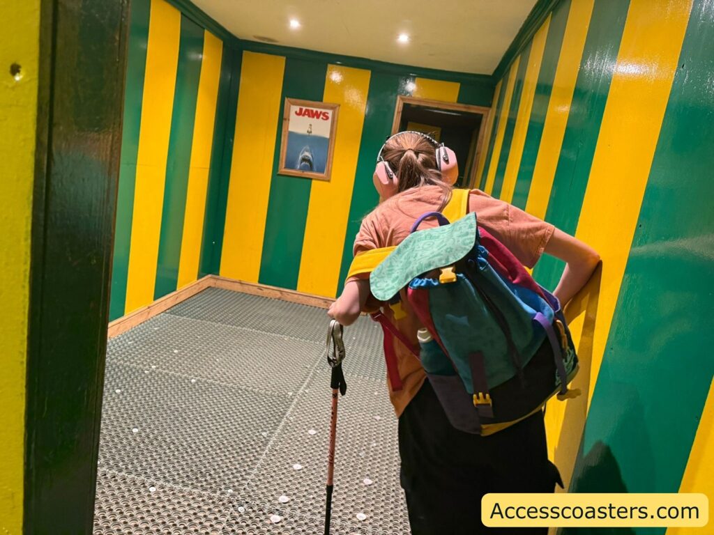 Iona with a colorful backpack and pink headphones walking through a room with green and yellow vertical striped walls. The person is using a walking stick  navigation and is touching the wall for support. The floor has a textured, metallic grid pattern