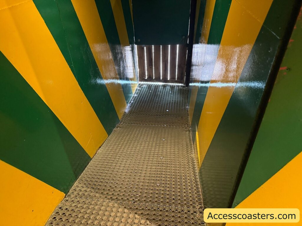 Narrow passageway with green and yellow diagonal stripes on the walls. The floor is made of a textured, perforated metal surface. At the end of the passage, there is a partially open gate 