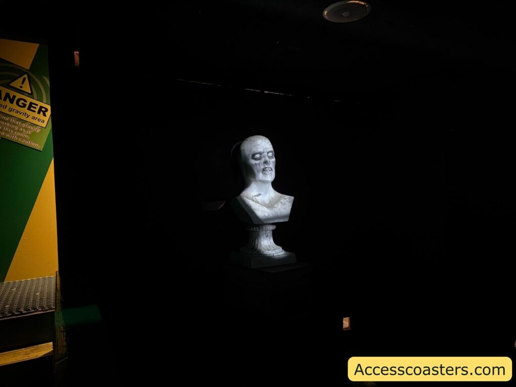 Marble bust in a dark room. The bust is illuminated and depicts a stern-looking man with a bald head and prominent facial features. To the left, there is a yellow and green warning sign that reads "DANGER: Altered gravity area" with additional smaller text below it. The background is mostly black, making the bust and the sign stand out.
