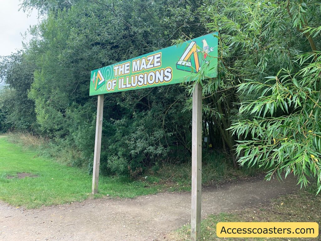 Entrance sign for an outdoor attraction called "The Maze of Illusions." The sign is mounted on two wooden posts and features colorful graphics, including a cartoonish triangular maze and playful illustrations. The path beneath the sign leads into a dense, green wooded area