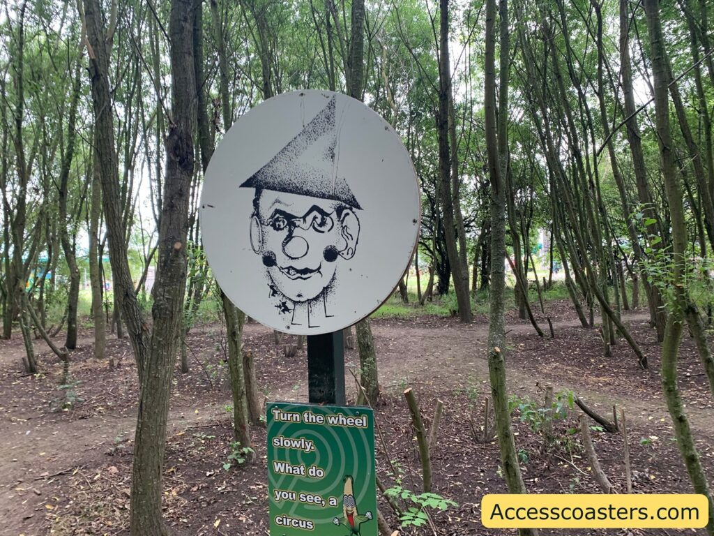 ircular sign with a black-and-white drawing of a clown's face, featuring a pointed hat, round nose, and exaggerated facial features, mounted on a post among thin trees in a wooded area. Below the sign, a green instruction panel reads: "Turn the wheel slowly. What do you see, a circus,"