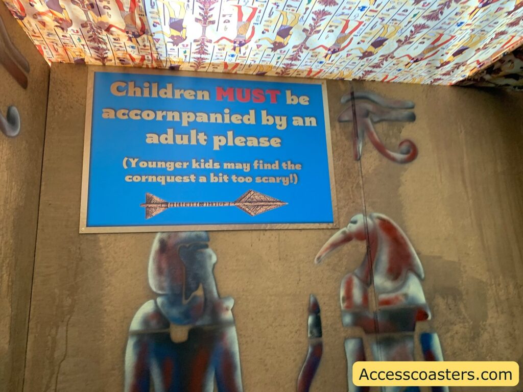 Blue sign with yellow and red text on a wall decorated with Egyptian-style art. The sign reads:

Children MUST be accompanied by an adult please
(Younger kids may find the cornquest a bit too scary!)

Below the sign, there are painted figures resembling ancient Egyptian gods. The ceiling is covered with a patterned fabric featuring hieroglyphic-like designs. 