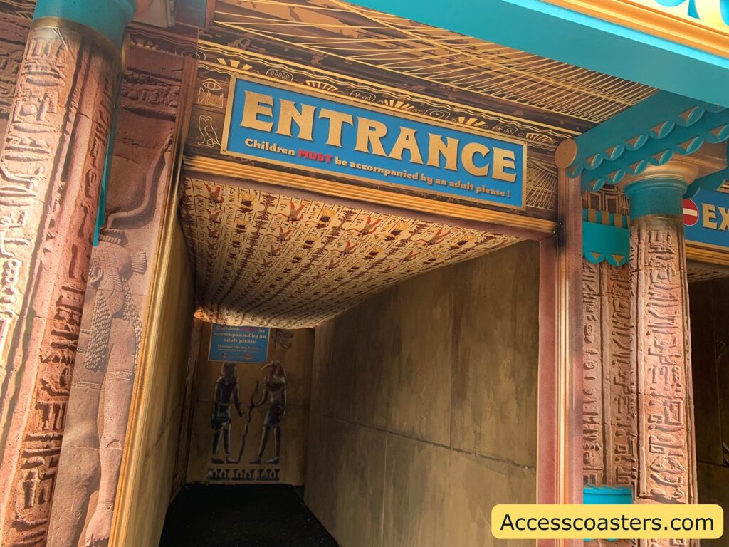 Egyptian-themed entrance to an attraction, with hieroglyphics and ancient-style artwork decorating the walls and ceiling. The entrance sign is blue with gold trim and reads "ENTRANCE" in large letters, with a smaller message underneath stating, "Children MUST be accompanied by an adult please!" 