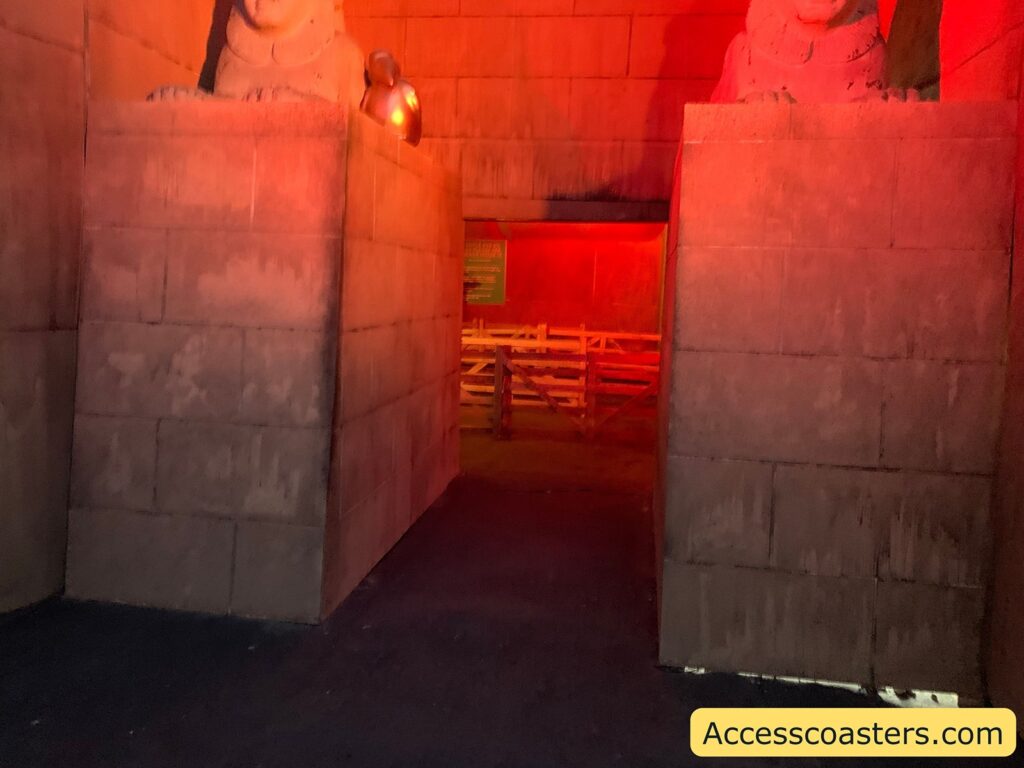 Dimly lit corridor with stone-like walls and statues. The scene is bathed in a reddish-orange light, giving it an ancient or mystical atmosphere. At the entrance, there are two large stone structures with what appear to be partial statues or carvings on top. Beyond the entrance, there is a wooden barrier or fence, and a green sign can be seen in the background.