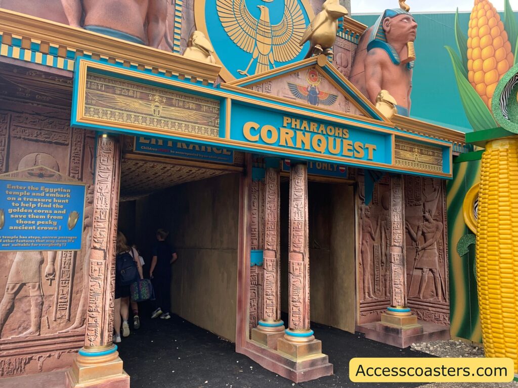 attraction entrance with an ancient Egyptian motif. The entrance is called "Pharaohs Cornquest," with large, bold blue and yellow letters above the doorway. The facade features Egyptian-style columns with hieroglyphics, statues of pharaohs, and a large golden winged scarab above the sign. To the right, there is a giant ear of corn, tying into the "corn" theme.