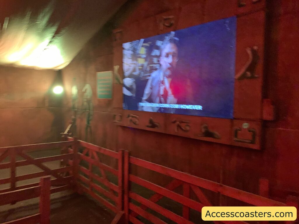 queue area with reddish-brown walls and wooden railings guiding visitors. On the wall, there is a large screen displaying a video of a man speaking, with English subtitles partially visible at the bottom. The screen is bordered by decorative symbols resembling ancient hieroglyphs or carvings.