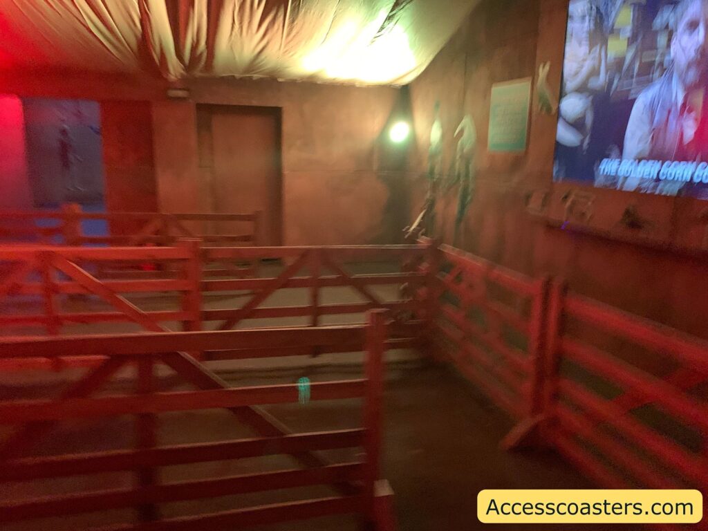 
Indoor amusement park or attraction queue area with wooden railings forming a zigzag path for guests to wait in line. The lighting is dim and warm, with red and yellow tones. The ceiling is draped with fabric, and there are some decorations on the wall, including what appear to be cornstalks. On the right wall, a large screen