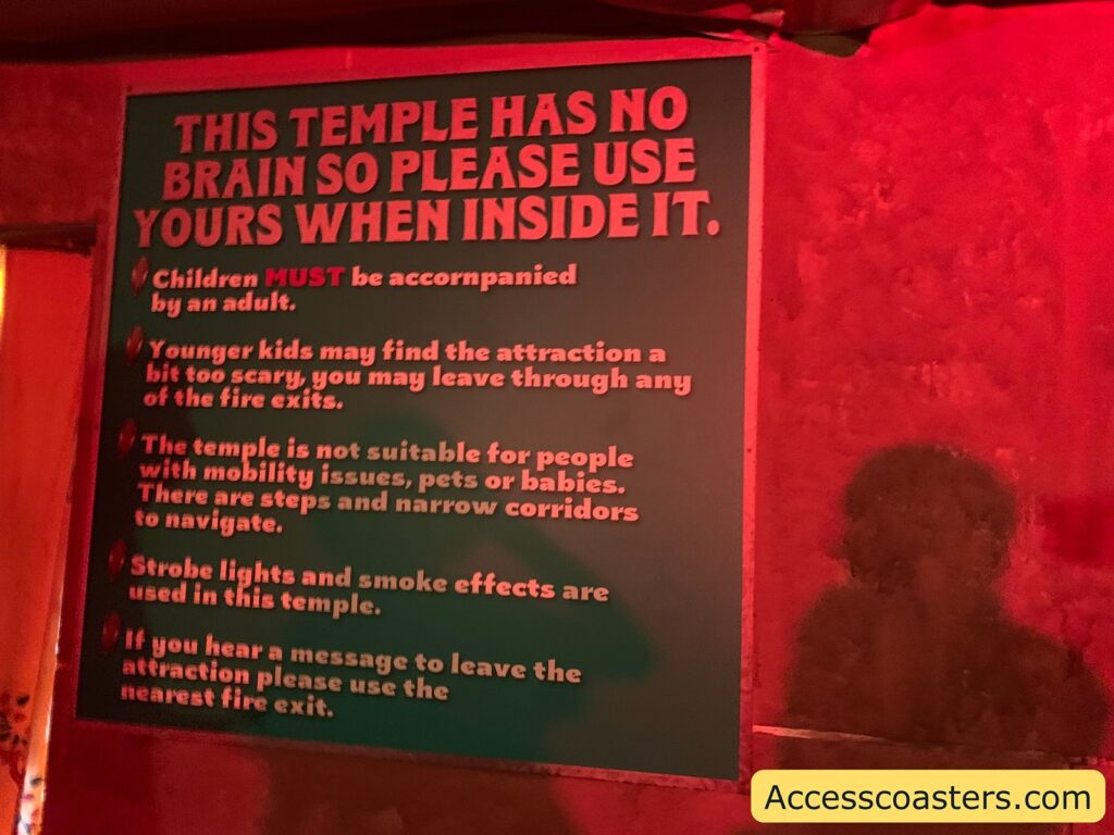 Large warning sign inside a dimly lit, red-toned area. The sign reads:

THIS TEMPLE HAS NO BRAIN SO PLEASE USE YOURS WHEN INSIDE IT.

- Children MUST be accompanied by an adult.
- Younger kids may find the attraction a bit too scary, you may leave through any of the fire exits.
- The temple is not suitable for people with mobility issues, pets or babies. There are steps and narrow corridors to navigate.
- Strobe lights and smoke effects are used in this temple.
- If you hear a message to leave the attraction please use the nearest fire exit.