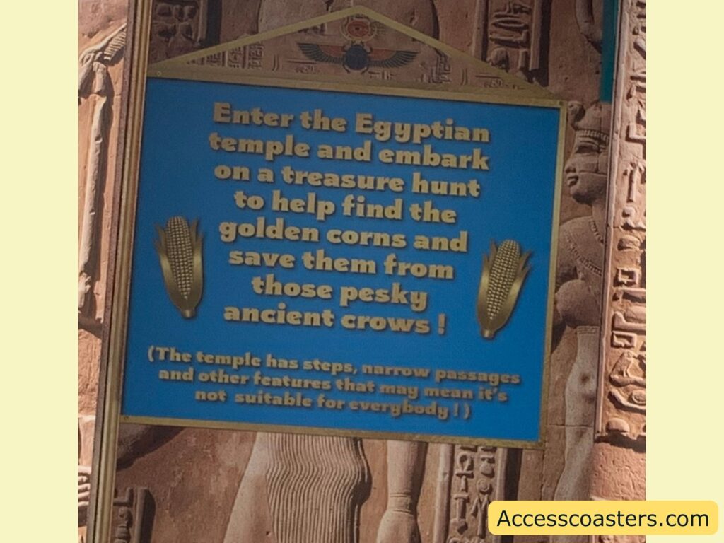 Blue and gold sign at an Egyptian-themed attraction. The sign reads:

"Enter the Egyptian temple and embark on a treasure hunt to help find the golden corns and save them from those pesky ancient crows!"

There are two golden corn illustrations on either side of the text. Below, in smaller text, it says:

"(The temple has steps, narrow passages and other features that may mean it's not suitable for everybody!)"

The background shows Egyptian-style carvings and statues. 