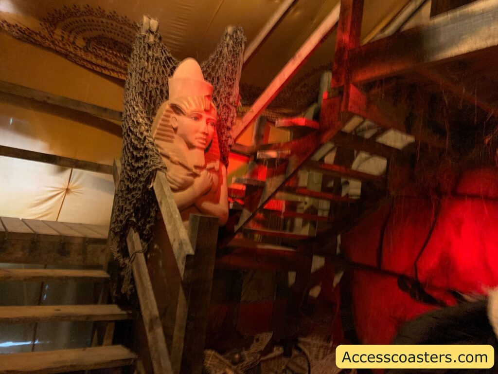 Dimly lit indoor staircase scene with an Egyptian pharaoh statue. The statue, resembling a traditional pharaoh with a headdress, is positioned at the base of a wooden staircase. The area is decorated with netting draped along the railings and illuminated by red lighting