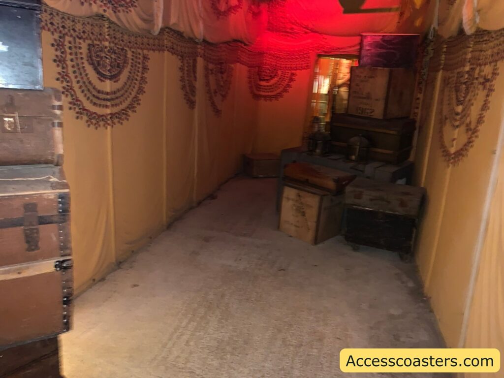 Dimly lit corridor with yellow draped fabric walls, decorated with maroon patterns resembling garlands or beads. The corridor is lined with several old-fashioned wooden and metal trunks and crates, stacked and scattered 