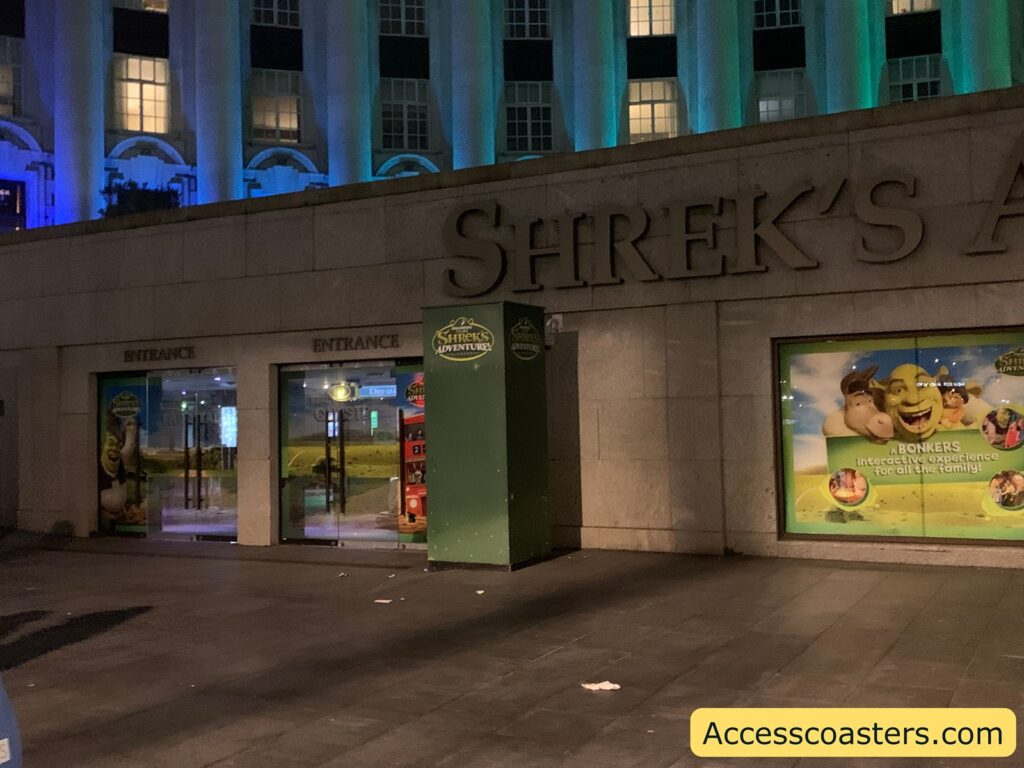 Entrance to Shrek's Adventure attraction at night. The image shows a building facade with large stone walls and tall windows above, illuminated by blue and green lights. The words "SHREK'S A" are visible in large letters on the wall, likely part of the full name "Shrek's Adventure." There are two glass entrance doors labeled "ENTRANCE" with Shrek-themed decorations. To the right, a large poster features Shrek, Donkey, and other characters, with the text: "A BONKERS interactive experience for all the family!"