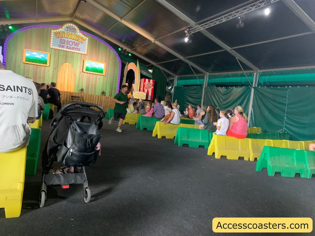 Indoor stage show setup with colorful plastic benches arranged in rows, mostly yellow and green. The stage at the front has a playful, carnival-like design with a large sign reading "THE CORNIEST SHOW ON EARTH" in bright lights, flanked by two screens displaying cartoonish images. the host with a micrphine in the audiance 
