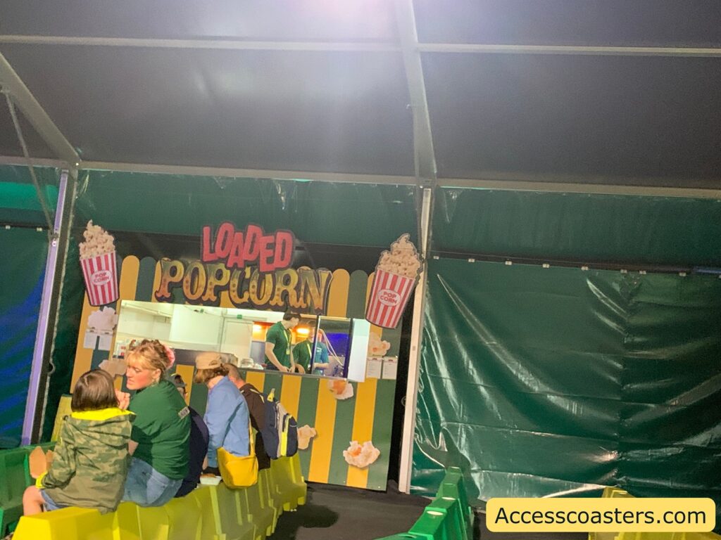 Popcorn concession stand scene. The image shows a stand with a sign reading "Loaded Popcorn" decorated with images of popcorn buckets. The stand is set up under a tent with green and yellow striped walls