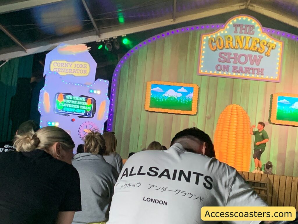 Indoor stage show with an audience. The stage has a large sign reading "THE CORNIEST SHOW ON EARTH" in colorful letters, with two screens framed by corn cobs showing a cartoon cornfield. A performer stands on stage near a large corn decoration. To the left, there’s a whimsical machine labeled "CORNY JOKE GENERATOR" with a screen that says, "WE THINK YOU'RE EVEN CLEVERER THAN CHAT-GPT!"