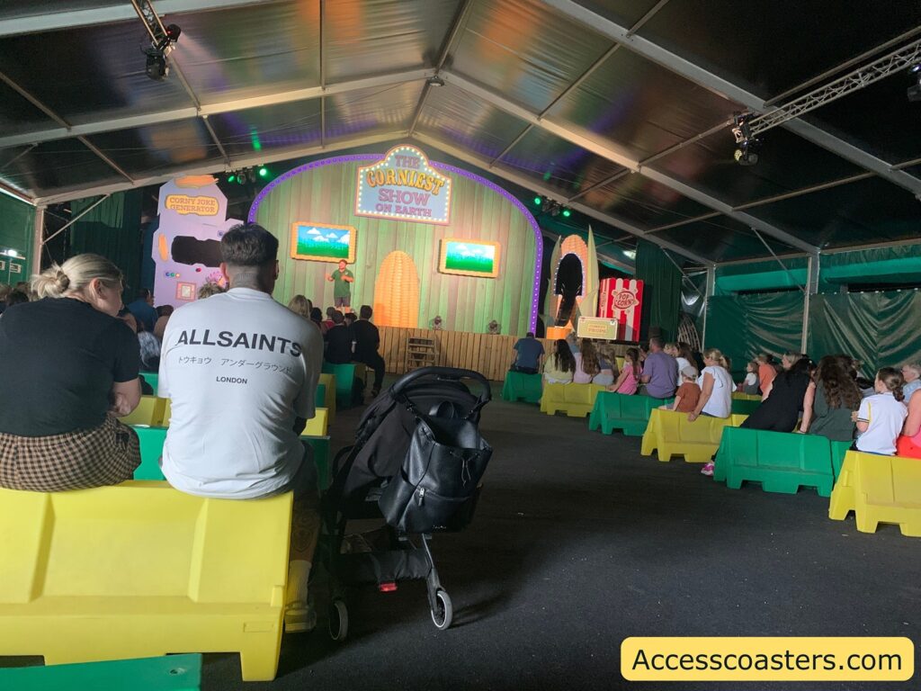 Indoor stage show with an audience seated on colorful plastic benches. The stage has a playful, carnival-like design with a large sign reading "The Corniest Show on Earth" and decorations including a corn cob and popcorn box. There are two screens on the stage, and a performer is speaking to the audience. 
