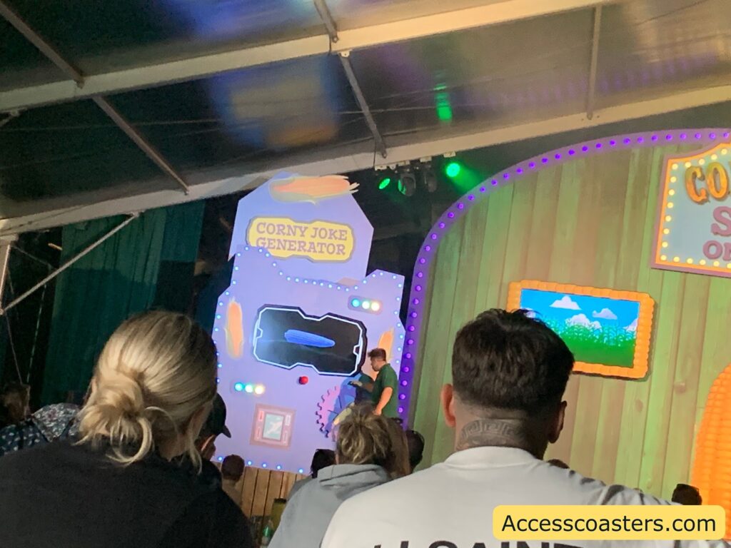 ndoor stage show scene. The photo shows an audience seated and watching a stage performance. On stage, there is a large, colorful prop labeled "CORNY JOKE GENERATOR" with lights and cartoonish decorations. A person stands near the prop, possibly a performer. 