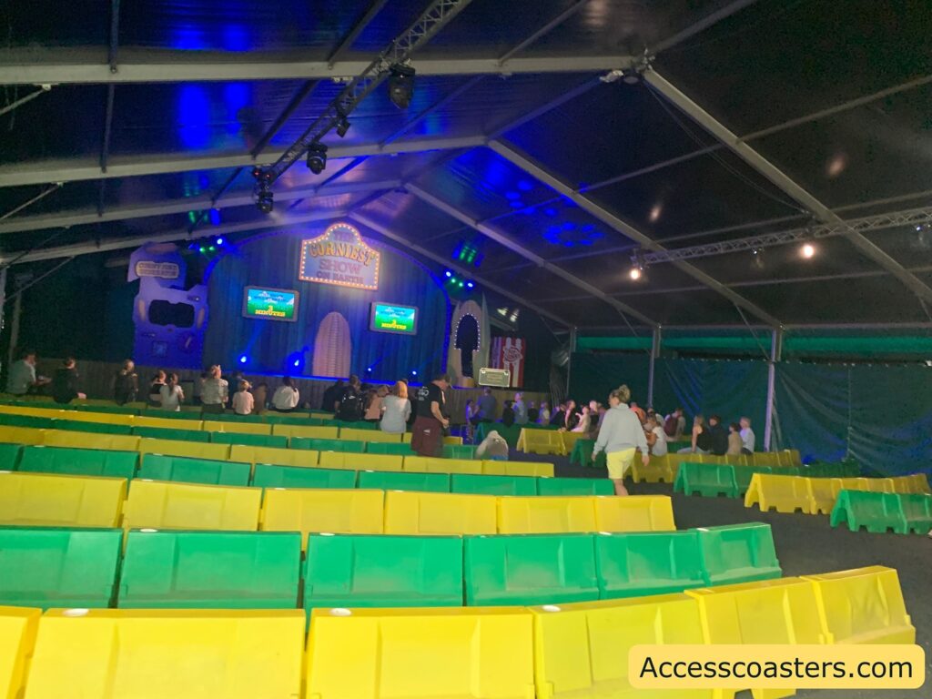 Indoor show venue with rows of yellow and green plastic benches, a stage at the front with a sign reading "Corniest Show," and two screens displaying the word "Next Show." The audience is sparsely seated, mostly towards the front. The venue is under a large tent structure with blue and purple stage lighting.