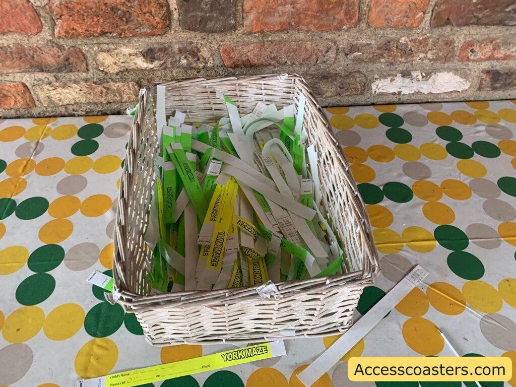 Wicker basket filled with paper wristbands, mostly white and some green, sitting on a table covered with a polka dot tablecloth in yellow, green, and beige. The wristbands have text on them, with some yellow ones labeled "YORKMAZE