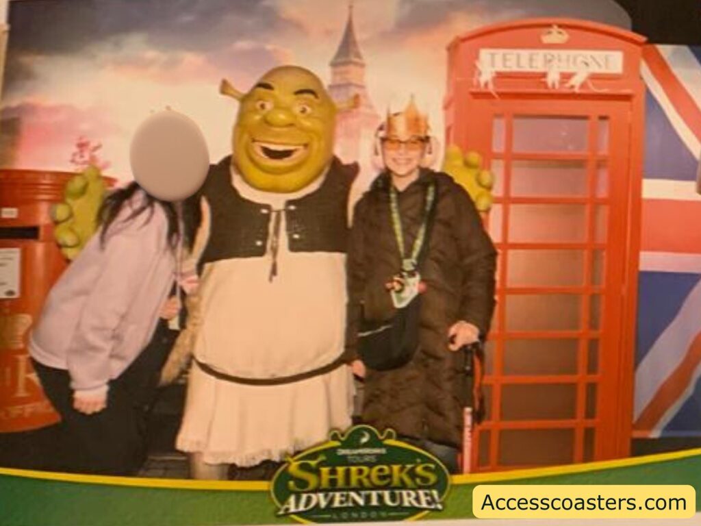 Theme park photo with Shrek. In the center, there is a person in a Shrek costume with green skin, a big smile, and wearing Shrek’s signature vest and tunic. On Shrek’s left, a person with long dark hair, wearing a light-colored jacket and black pants, is leaning in; their face is blurred for privacy. On Shrek’s right, another person is standing, wearing a dark puffy coat, a crown, glasses, and a lanyard around their neck, holding a walking stick. Behind them is a bright red British telephone box and a partial view of the Union Jack flag. 