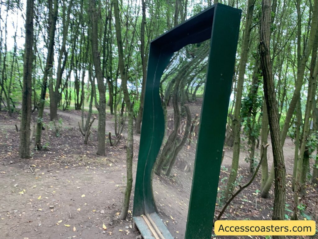 Distorted funhouse mirror in a wooded area. The mirror is tall, rectangular, and framed in dark green, with a wavy surface that bends and twists the reflection of the surrounding trees and ground, creating a playful, warped effect. 