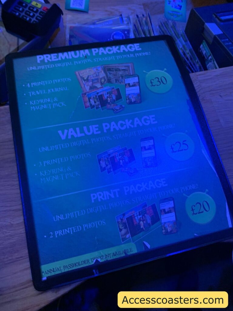 Price list for photo packages at an amusement park or attraction, displayed under blue lighting on a wooden counter. There are three packages shown:

Premium Package (£30): Includes unlimited digital photos sent straight to your phone, 4 printed photos, a travel journal, and a keyring & magnet pack.
Value Package (£25): Includes unlimited digital photos sent straight to your phone, 3 printed photos, and a keyring & magnet pack.
Print Package (£20): Includes unlimited digital photos sent straight to your phone and 2 printed photos.
At the bottom, there is a note: "Annual passholder discount available."