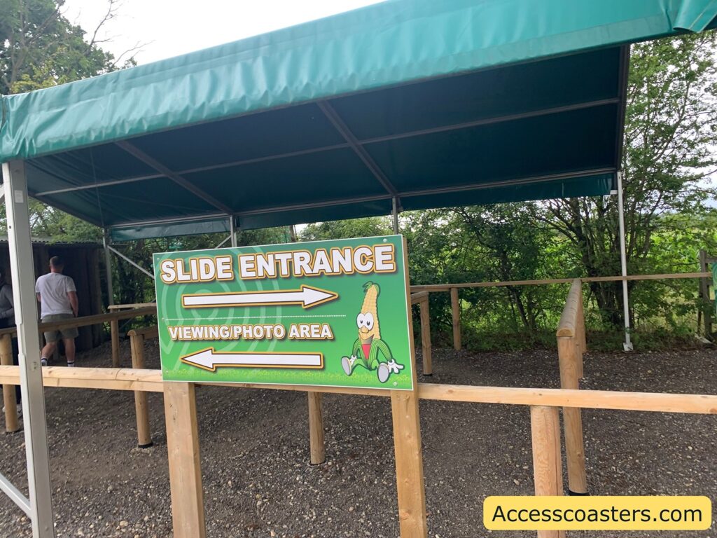 Outdoor queue area with a green canopy overhead and wooden railings. A large green sign in the foreground reads "SLIDE ENTRANCE" with a right-pointing arrow, and "VIEWING/PHOTO AREA" with a left-pointing arrow. The sign features a cartoon corn character sitting and smiling. In the background,