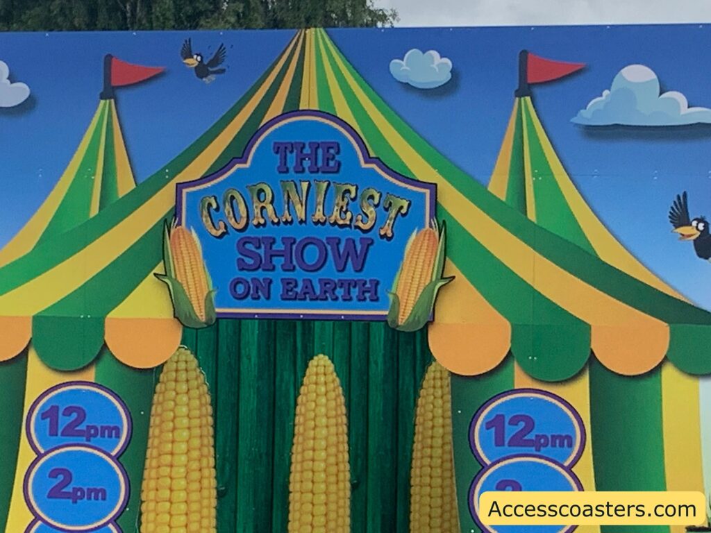 Colorful carnival-themed entrance display with a circus tent design in green, yellow, and orange. The sign at the center reads "THE CORNIEST SHOW ON EARTH" with images of corn cobs on either side. Below the sign, there are two large corn cobs and circular signs on both sides listing showtimes: "12 pm" and "2 pm." Cartoon birds and clouds decorate the top.