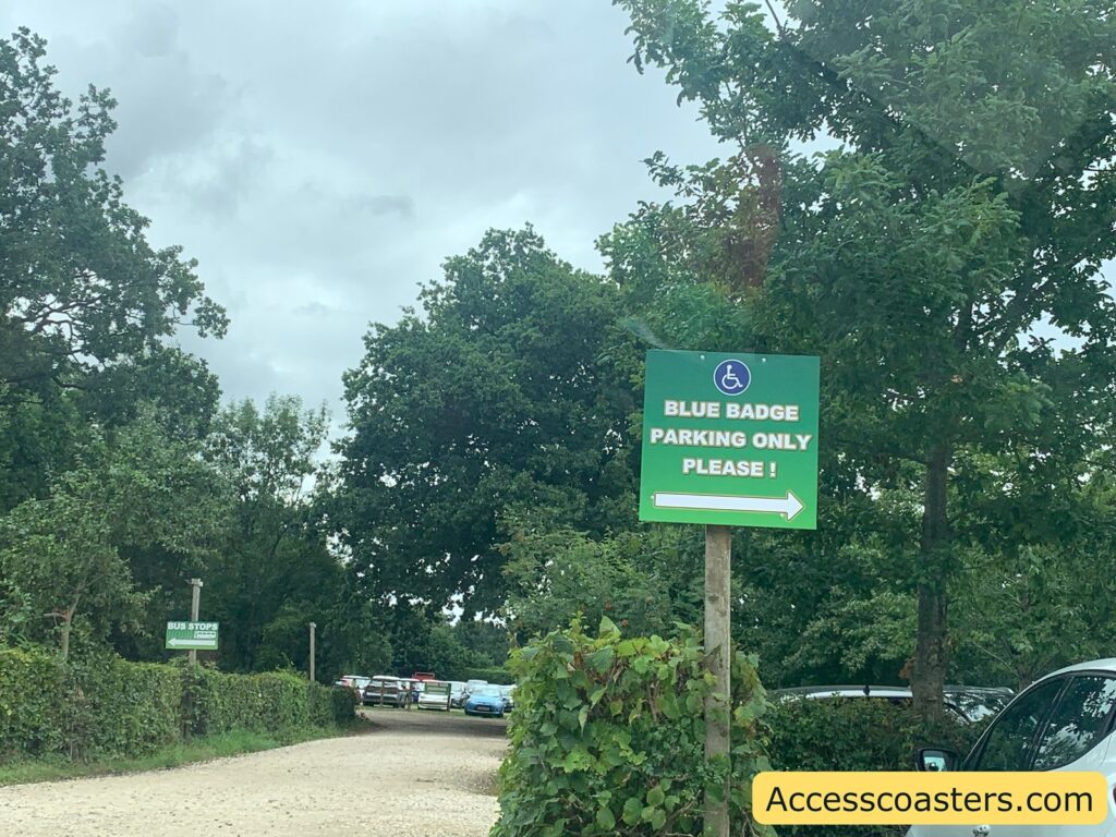 Parking area with a green sign that reads "BLUE BADGE PARKING ONLY PLEASE!" and features a wheelchair accessibility symbol. The sign has a white arrow pointing to the right, indicating the direction for accessible parking. There are several cars parked in the background along a gravel road, surrounded by green trees and bushes.