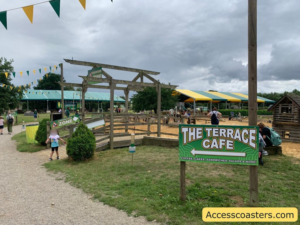 Outdoor play area at a family attraction, with wooden structures, sand, and covered seating areas. There are colorful yellow and green pennant banners strung overhead. A large sign in the foreground reads "THE TERRACE CAFE" with an arrow pointing left, advertising coffee, cakes, sandwiches, salads, and more. Another sign points to a "CONSTRUCTION ZONE" play area.
