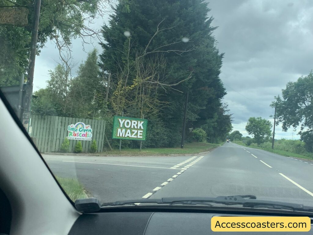 Roadside view from inside a car, showing a sign for "YORK MAZE" on a green fence to the left, next to another colorful sign for "Little Green Rascals Children's Organic Day Nursery." The road is bordered by tall trees and greenery, with a cloudy sky above. The dashboard and part of the windshield of the car are visible in the foreground.