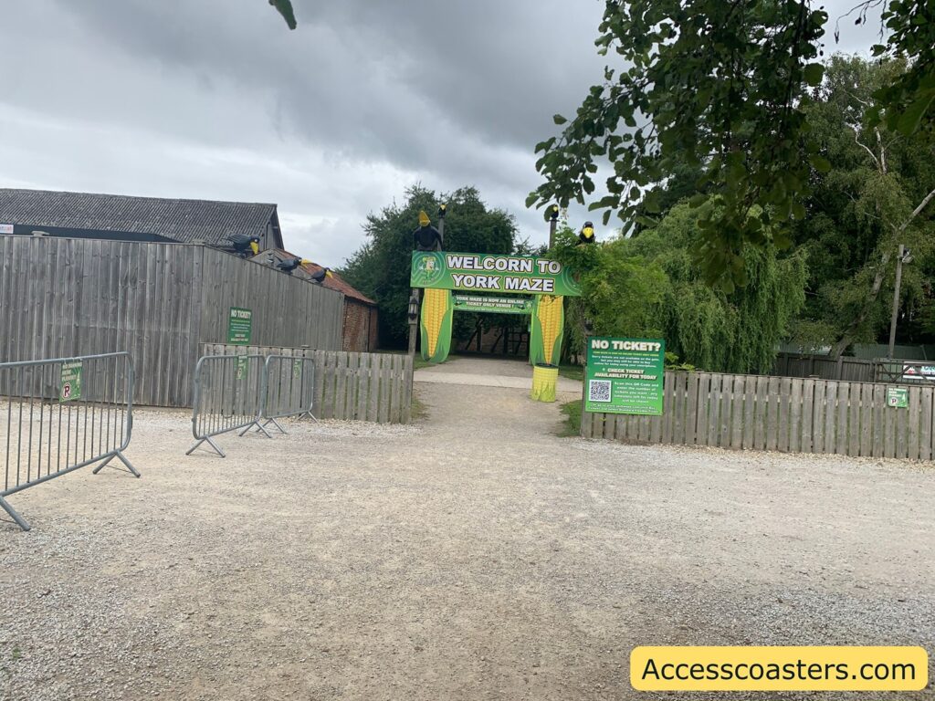Entrance to York Maze. The image shows a wide gravel path leading up to a green and yellow entrance archway decorated with corn cobs and cartoon faces. The sign above the entrance reads "WELCOME TO YORK MAZE." There are metal barriers and wooden fences guiding visitors towards the entrance. Several green signs provide information, including one that says "NO TICKET?" with instructions and a QR code for checking ticket availability. 