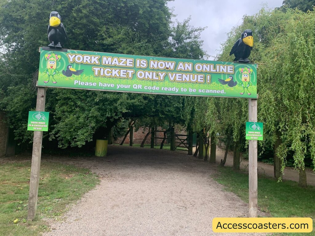 Entrance archway to an outdoor venue, with a large green sign overhead that reads: "YORK MAZE IS NOW AN ONLINE TICKET ONLY VENUE! Please have your QR code ready to be scanned." The sign features cartoon images of a corn character and black birds with yellow beaks. On top of the sign, there are two large black bird statues, one on each side. Smaller green signs on the wooden posts say, "YORK MAZE IS A CASHLESS VENUE"