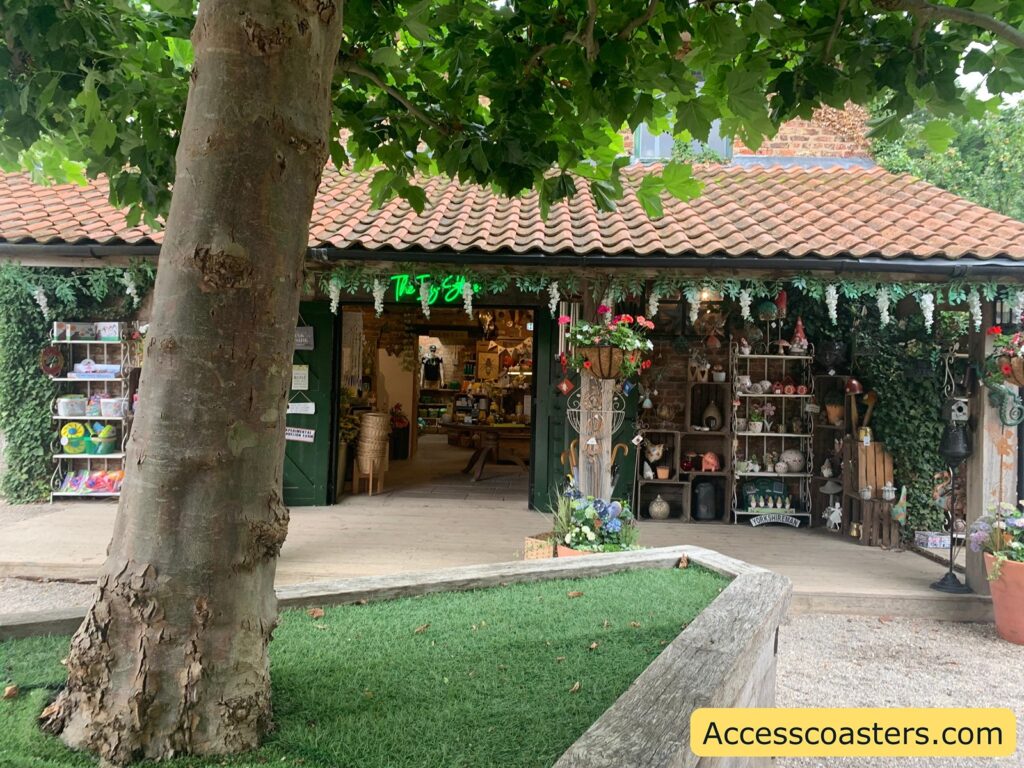 Outdoor garden shop scene. There is a large tree in the foreground with a thick trunk and leafy branches providing shade. Behind the tree, there is a rustic shop with a red-tiled roof and green doors, partially open to reveal the interior filled with various items. The shop's entrance is decorated with hanging plants and flowers, and shelves on either side display garden ornaments, pots, and tools. The shop sign above the door is written in green neon letters. In front of the shop, there is a raised bed with artificial grass bordered by wooden planks. 