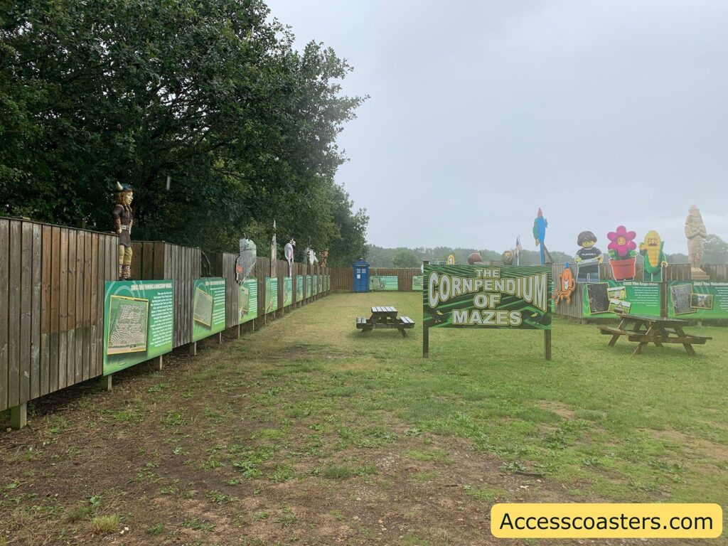 Outdoor attraction area with a large green sign in the center reading "The Cornpendium of Mazes." The area is grassy with a few wooden picnic tables scattered around. Along the left side, there is a wooden fence with green informational boards and various character statues on top, including a Viking and other figures. In the background, there are more colorful, cartoon-like character statues,