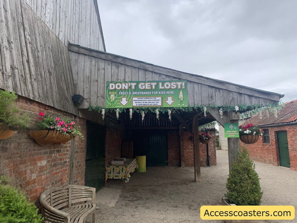 Covered entrance area at a rustic outdoor venue, with a large green and white sign overhead that reads "DON'T GET LOST! FREE I.D. WRISTBANDS FOR KIDS HERE." The sign features cartoon corn characters and arrows pointing downward. Below the sign, there is a table covered with a patterned cloth and wristband 