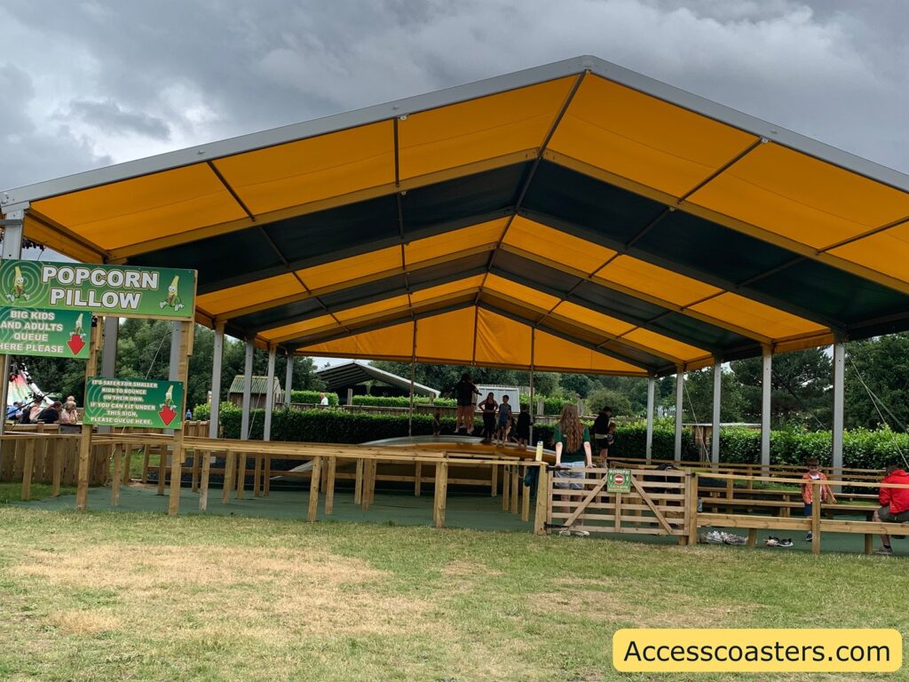 Outdoor play area with a large yellow and green canopy covering a wooden fenced structure. Inside, there is an inflated bouncy surface called the "Popcorn Pillow," where several children and adults are playing. Signs indicate separate queues for big kids/adults and smaller kids. The ground is grassy outside the fenced area, and there are shoes left near the entrance. 
