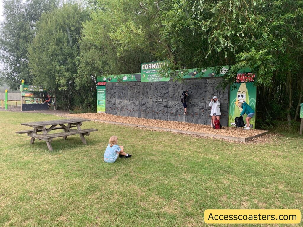 Outdoor playground area with a small artificial climbing wall. The wall has gray, rock-like holds and is set against a backdrop of green trees. a sign on the climbing wall that reads "CORNWALL" and features a cartoon corn character. 