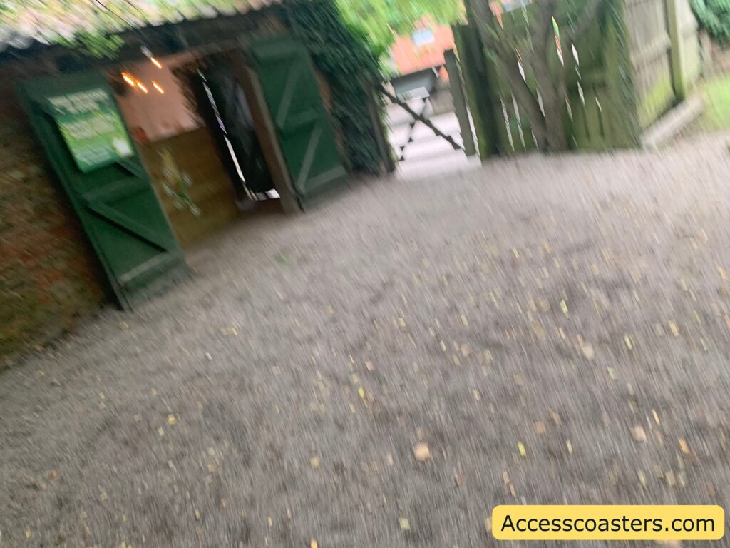 Outdoor scene with a gravel pathway, a rustic building with green double doors open, and a wooden gate in the background. The image is slightly blurry. There is some greenery around the area, and a sign is visible on the left door, but the text is not clear due to the blur. 