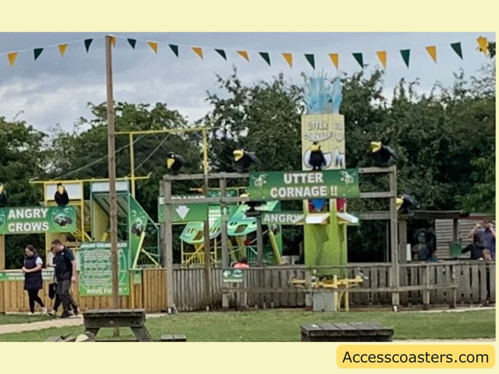 Outdoor amusement park area with a playful theme. There are large, cartoonish black birds with yellow beaks perched on a wooden structure. Signs read "UTTER CORNAGE!!" and "ANGRY CROWS," suggesting a pun on "utter carnage" and the popular game "Angry Birds." The area is decorated with green and yellow flags