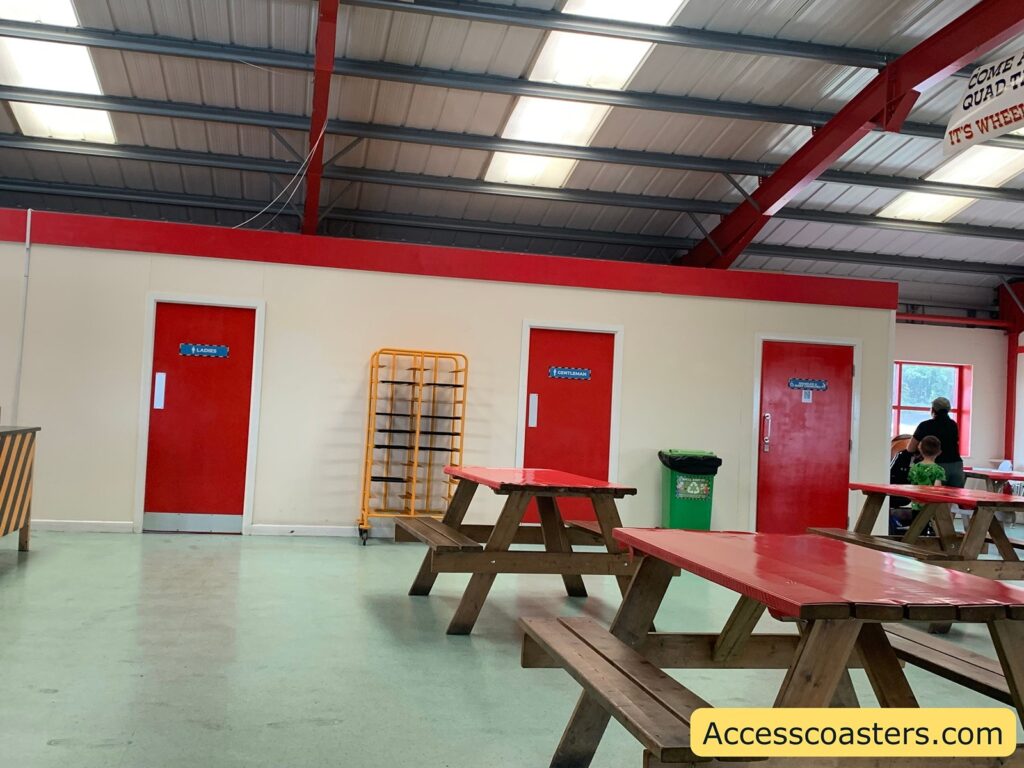 Indoor cafeteria setting with picnic-style wooden tables that have bright red tops. The background wall is beige with a red border at the top and has three red doors. Above each door are blue signs: the left door says "LADIES", the middle door says "GENTS/MALE",