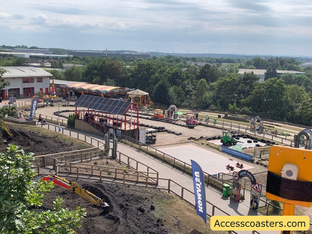 Outdoor amusement park area featuring a dirt digging zone with a yellow excavator, winding pathways, and various ride attractions a door bell is visable in the right corner to press for help