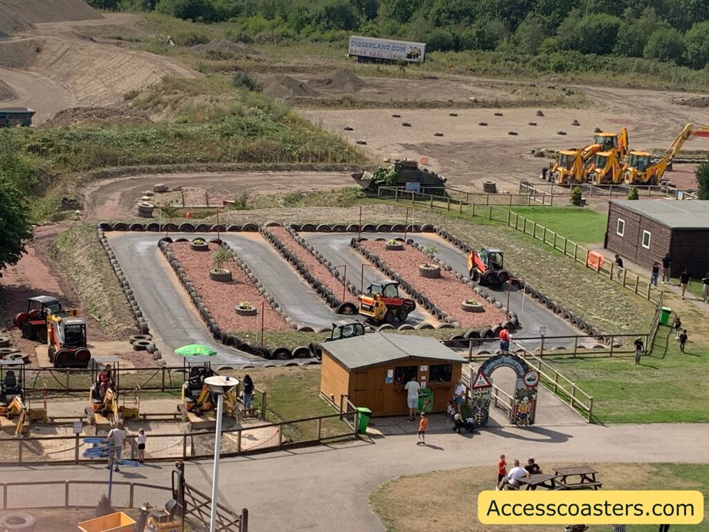 Amusement park attraction with a construction theme. The main feature is a small go-kart-style track with several miniature construction vehicles, such as rollers and diggers, being driven by children.